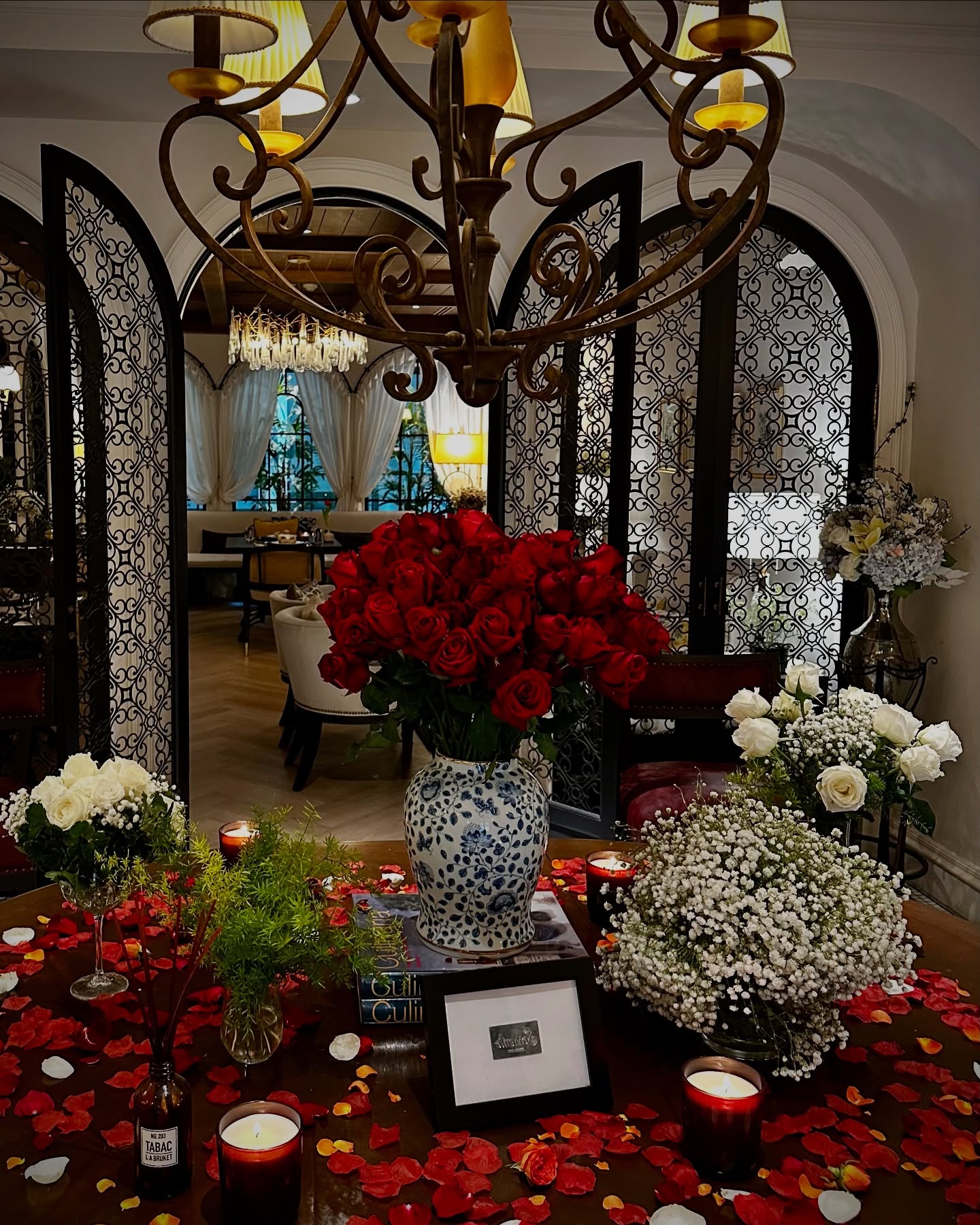 Luxurious and romantic restaurant interior featuring a large bouquet of red roses, scattered rose petals, and warm candlelight under an ornate chandelier.